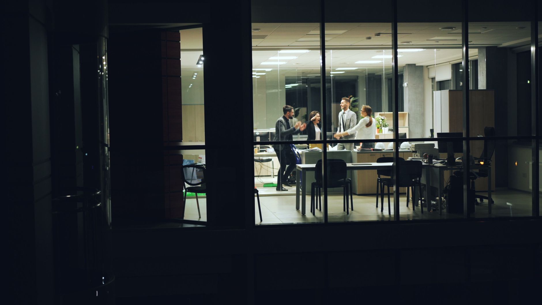 A group of five professionals engaging and celebrating in a modern office space during nighttime, visible through large glass windows.