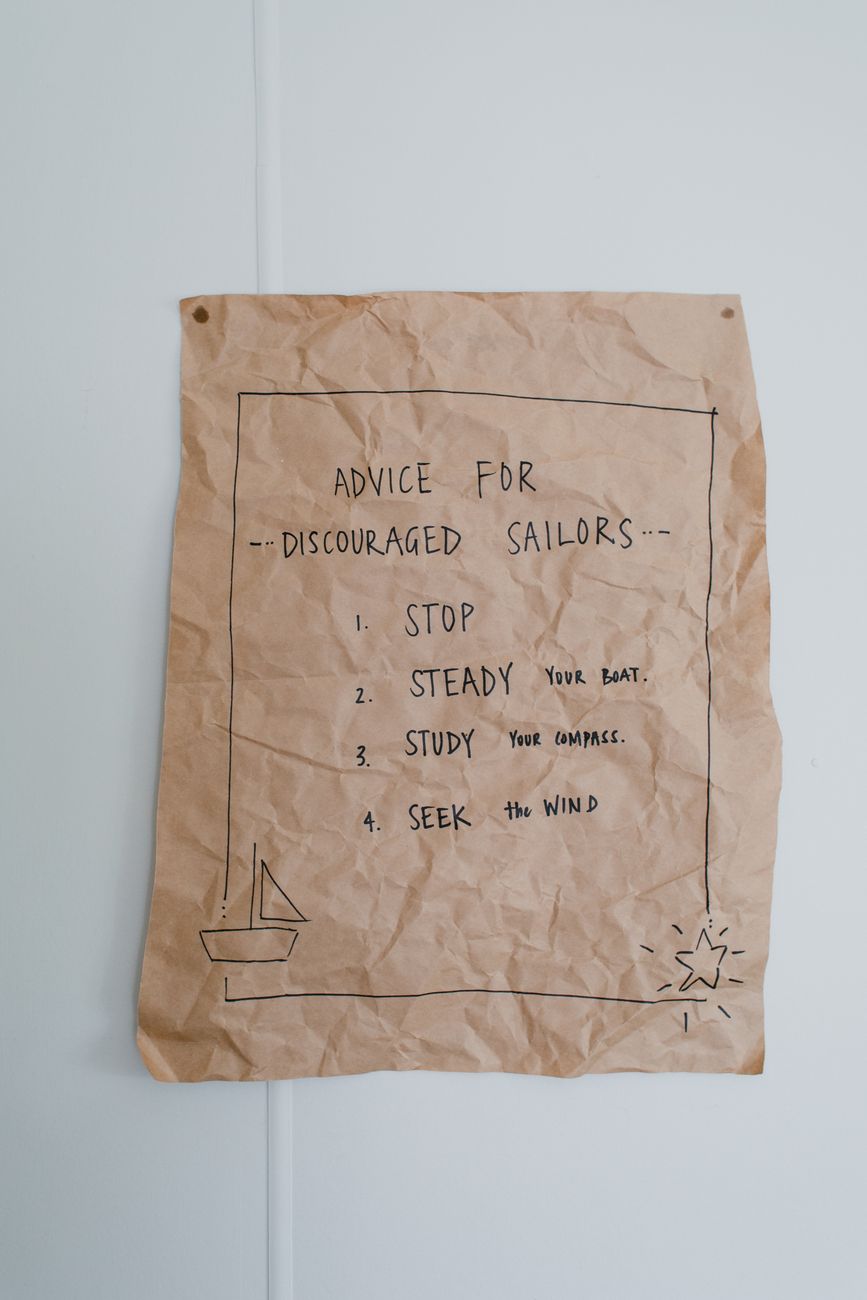 A wrinkled brown paper with handwritten advice for discouraged sailors, listing four steps: 1. STOP, 2. STEADY your boat, 3. STUDY your compass, 4. SEEK the wind. Illustrated with a small sailboat and decorative elements.